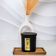 Load image into Gallery viewer, A matte black Ocean Breeze candle jar with a gold foil label sits centered on a wooden tray in front of a modern white ceramic vase filled with dried pampas grass. The background features soft white wave textures for a serene, minimal look.