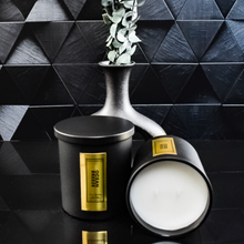 Load image into Gallery viewer, A pair of Ocean Breeze candles are displayed on a black reflective surface. One candle is upright, while the other lies on its side showing the clean white wax. The setup includes a sculptural matte black vase with eucalyptus for a bold, elegant composition.
