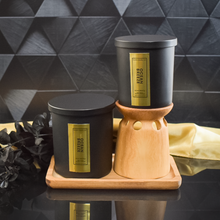 Load image into Gallery viewer, Two Ocean Breeze candles in black jars with vertical gold labels are displayed on a wooden tray. One is elevated on a wood candle riser. The black and gold background and dried botanicals create a moody, upscale vibe.