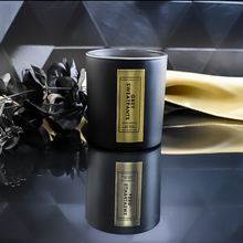 Load image into Gallery viewer, An open Grey Sweatpants candle in a matte black jar with a rich gold label. It sits on a reflective backgrounds. A gold satin cloth and black leaves enhance the candle’s luxurious presentation.