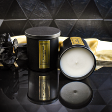 Load image into Gallery viewer, A pair of Grey Sweatpants candles are displayed on a black reflective surface. One candle is upright, while the other lies on its side showing the clean white wax. The setup includes a sculptural matte black vase with eucalyptus for a bold, elegant composition.