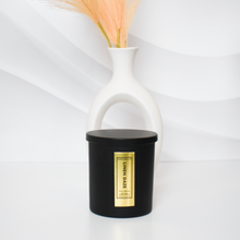 Load image into Gallery viewer, Stylish Linen Daze candle in a black matte jar with gold foil label, paired with a modern white vase and peach-toned dried florals for a clean aesthetic.