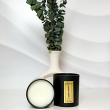 Load image into Gallery viewer, Handcrafted Linen Daze candle duo featuring creamy soy wax and matte black jars with gold foil labels, styled against a wave-textured white backdrop with fresh eucalyptus in a modern vase.