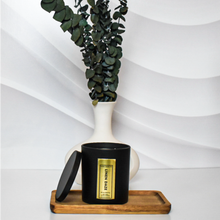 Load image into Gallery viewer, Matte black Linen Daze candle with gold foil label displayed on a wooden tray, paired with a modern white vase and lush eucalyptus stems, set against a sculpted white background for a clean, minimalist aesthetic.