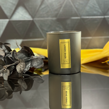 Load image into Gallery viewer, Luxury 12 oz Linen Daze candle in matte black jar with reflective gold foil label, elegantly staged against a diamond-textured black tile backdrop.