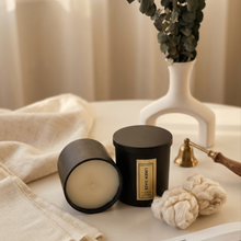 Load image into Gallery viewer, Two black candles on a surface with a plant and decorative items in the background.