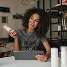 Load image into Gallery viewer, Woman sitting at a desk holding a phone and beauty product, wearing a 'Energy Protected' shirt.
