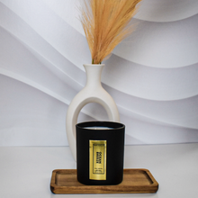 Load image into Gallery viewer, A matte black Ocean Breeze candle jar with a gold foil label sits centered on a wooden tray in front of a modern white ceramic vase filled with dried pampas grass. The background features soft white wave textures for a serene, minimal look.
