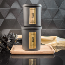 Load image into Gallery viewer, Two stacked black candle jars labeled Ocean Breeze with gold foil accents sit atop a wooden tray, styled against a geometric black tile backdrop with a soft gold satin drape and black foliage for a luxe, layered presentation.
