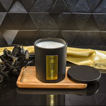 Load image into Gallery viewer, An open Ocean Breeze candle reveals its double wick inside a matte black jar with a rich gold label. It sits on a wooden tray with its lid beside it. A gold satin cloth and black leaves enhance the candle’s luxurious presentation.

