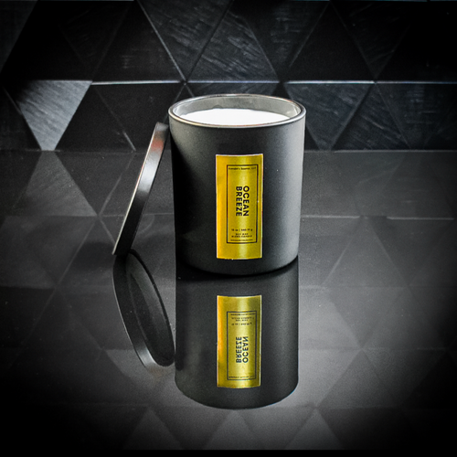 A sleek black candle jar labeled “Ocean Breeze” with a reflective gold foil label, displayed on a glossy black tiled surface. The candle lid is propped open, revealing the white wax inside, and the label reads “Kandie’s Scents, LLC” with minimalistic luxury branding.