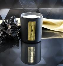 Load image into Gallery viewer, An open Grey Sweatpants candle reveals its single wick inside a matte black jar with a rich gold label. A gold satin cloth and black leaves enhance the candle’s luxurious presentation.
