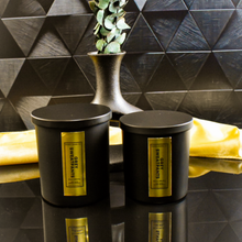 Load image into Gallery viewer, Two Grey Sweatpants candles in black jars with vertical gold labels are displayed on a reflective background. The black and gold background and dried botanicals create a moody, upscale vibe.

