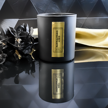 Load image into Gallery viewer, An open Grey Sweatpants candle inside a matte black jar with a rich gold label. A gold satin cloth and black leaves enhance the candle’s luxurious presentation.
