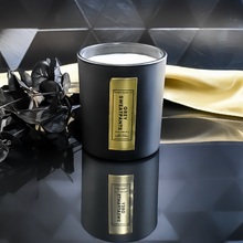 Load image into Gallery viewer, An open Grey Sweatpants candle reveals its double wick inside a matte black jar with a rich gold label. It sits on a reflective backgrounds.  A gold satin cloth and black leaves enhance the candle’s luxurious presentation.
