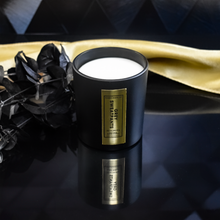 Load image into Gallery viewer, An open Grey Sweatpants candle in a matte black jar with a rich gold label. It sits on a reflective backgrounds.  A gold satin cloth and black leaves enhance the candle’s luxurious presentation.
