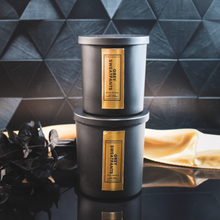 Load image into Gallery viewer, Two stacked black candle jars labeled Grey Sweatpants with gold foil accents sit on a reflective surface, styled against a geometric black tile backdrop with a soft gold satin drape and black foliage for a luxe, layered presentation.
