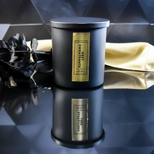 Load image into Gallery viewer, A closed Grey Sweatpants candle in a matte black jar with a rich gold label. A gold satin cloth and black leaves enhance the candle’s luxurious presentation.
