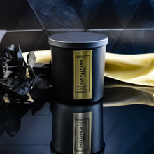 Load image into Gallery viewer, A closed Grey Sweatpants candle in a matte black jar with a rich gold label. A gold satin cloth and black leaves enhance the candle’s luxurious presentation.
