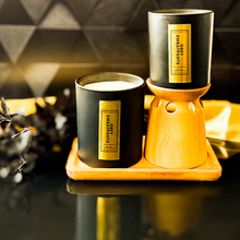 Load image into Gallery viewer, Two Grey Sweatpants candles in black jars with vertical gold labels are displayed on a wooden tray. One is elevated on a wood candle riser. The black and gold background and dried botanicals create a moody, upscale vibe.
