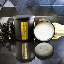 Load image into Gallery viewer, A pair of Grey Sweatpants candles are displayed on a black reflective surface. One candle is upright, while the other lies on its side showing the clean white wax. 
