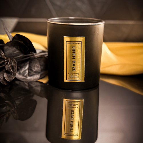 uxury Linen Daze candle in 8.5 oz matte black jar with gold foil label, reflected on a glossy black surface with dried eucalyptus and a golden accent cloth in the background.