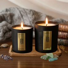 Load image into Gallery viewer, Two lit candles from 'Mom's Bakery' on a wooden tray with books and decorative stones in the background.
