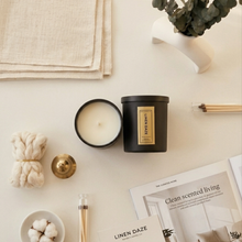 Load image into Gallery viewer, Two candles on a table with decorative items including a plant, newspaper, and cotton balls.
