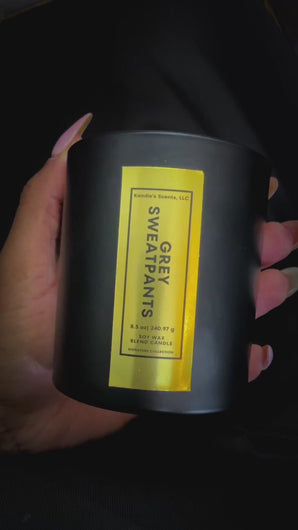 A close-up video of an 8.5 oz Grey Sweatpants candle held in one hand and gently rotated to highlight its elegant gold foil label. The candle is housed in a matte black jar, and the reflective label catches the light, showcasing the luxury and craftsmanship of Kandie’s Scents’ signature scent.
